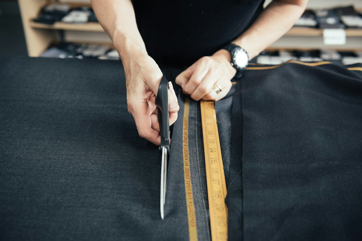 A tailor cutting dark suiting cloth with shears beside a tape measure